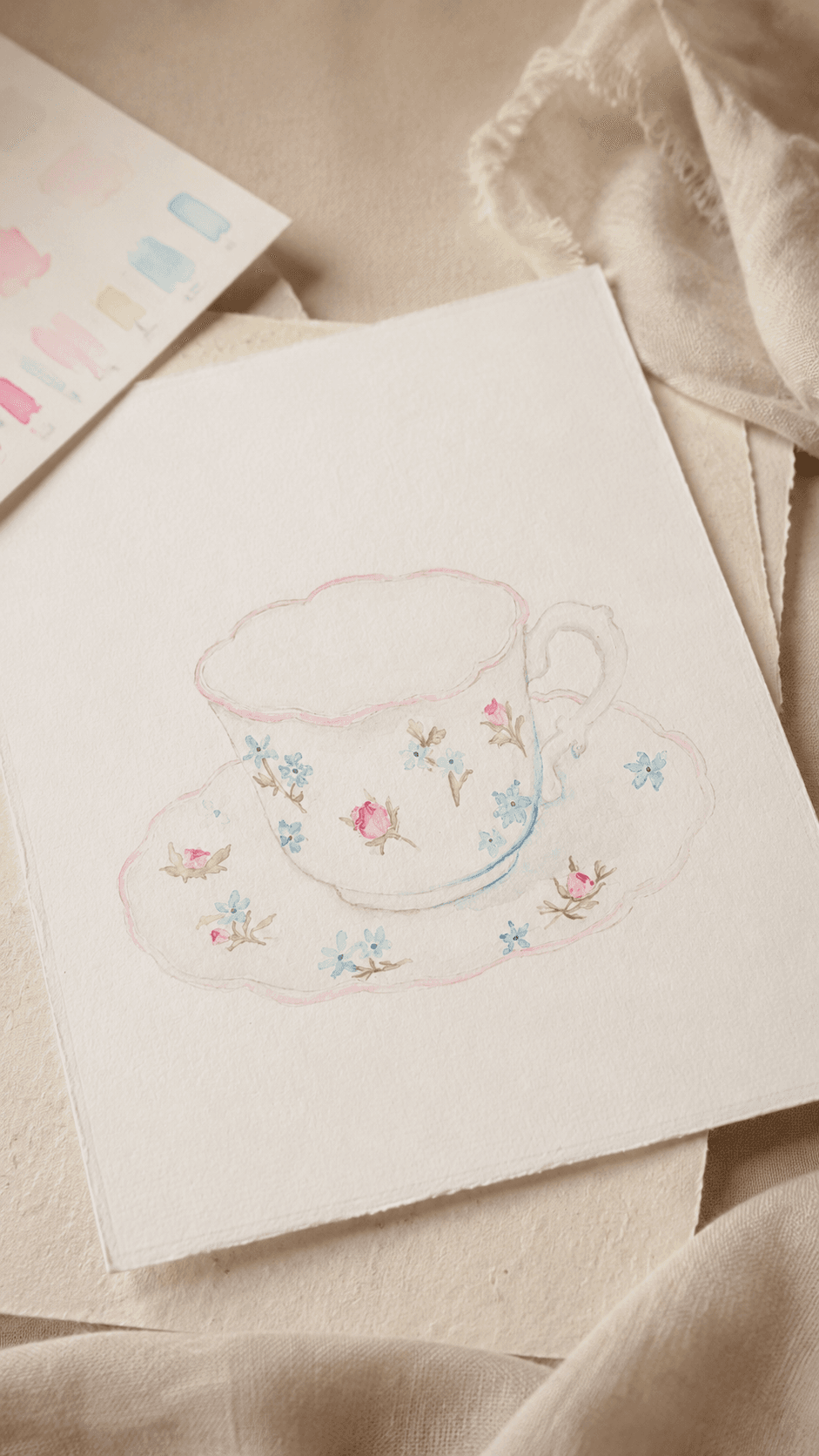 Watercolor painting of a scalloped teacup and saucer scattered with pink rosebuds and blue forget-me-nots, resting on soft linen beside color swatches.
