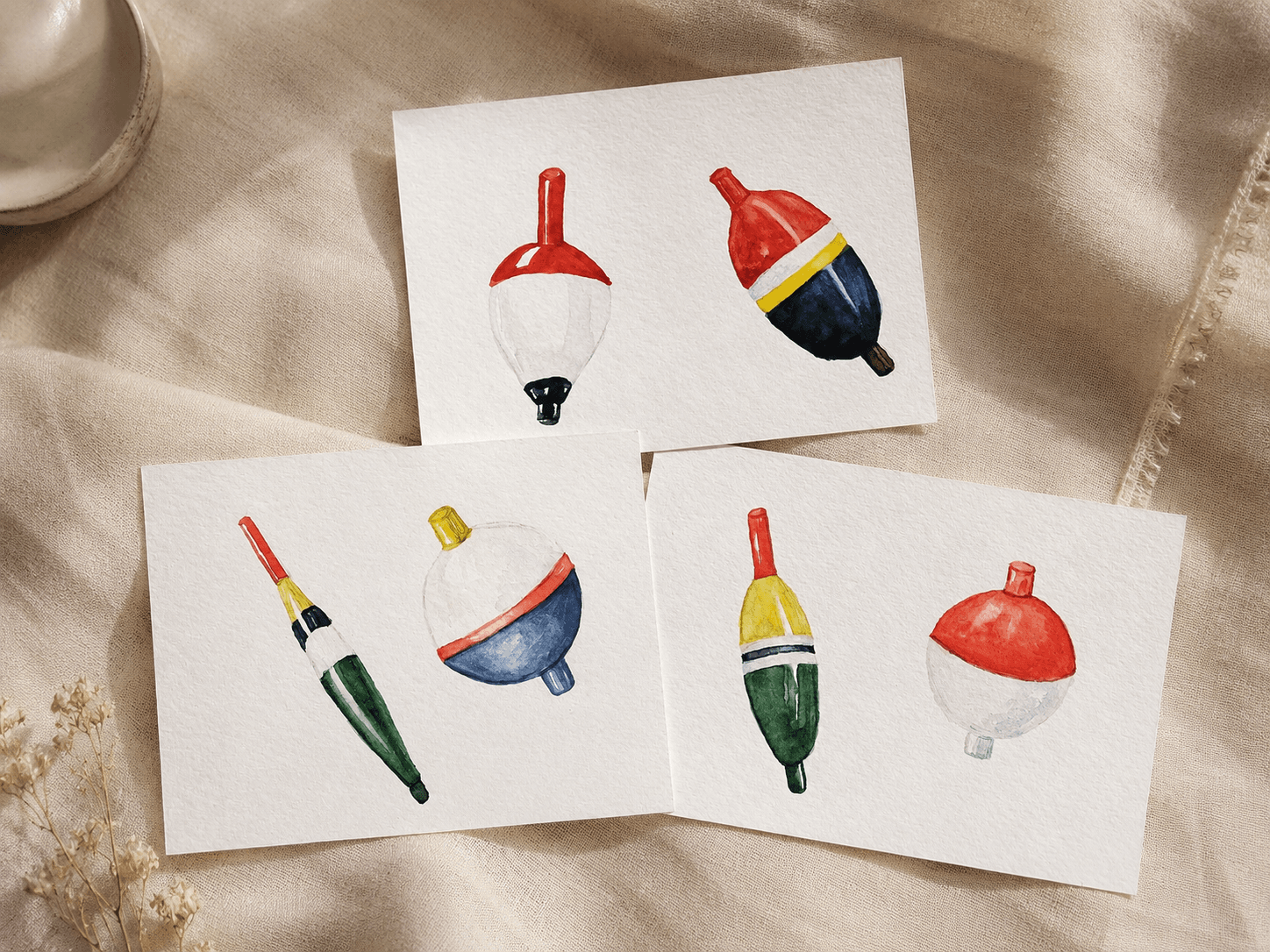 Set of three watercolor studies of vintage fishing bobbers in red, navy, green, and yellow, laid out on soft cream linen beside dried flowers.
